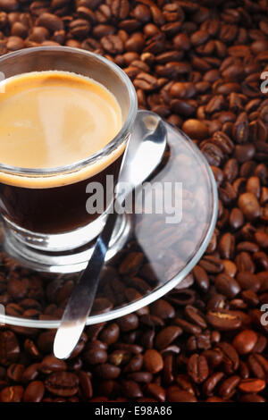 Eine Tasse schwarzen Kaffee in einer Schale Klarglas, mit passender Untertasse und Löffel aus Edelstahl, ruht auf einem Hintergrund von Kaffeebohnen Stockfoto