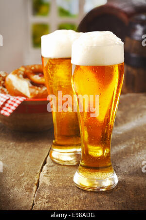 Zwei große Gläser golden Ale oder Bier mit einem guten Kopf des weißen Schaum auf einem alten hölzernen Küchentisch mit einem Korb mit Brötchen hinter Stockfoto