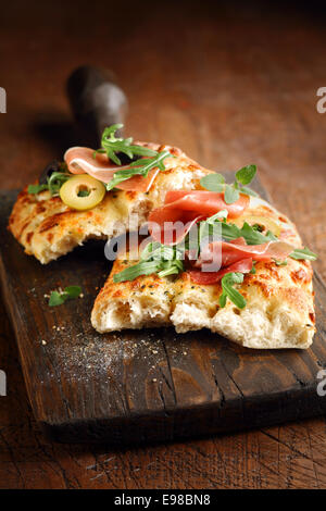 Frisch gebackene traditionelle herzhafte italienische Focaccia Brot mit Schinken und Oliven belegt und garniert mit Rucola auf eine alte Grunge geknackt Schneidbrett aus Holz Stockfoto