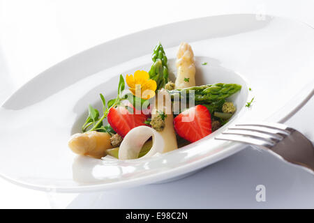 Leckeren Spargel Vorspeise mit kalten frischen grünen und weißen Spitzen gemischt mit Erdbeeren in eine schlichte weiße Keramikschale serviert und garniert mit eine kleine gelbe Blume Stockfoto