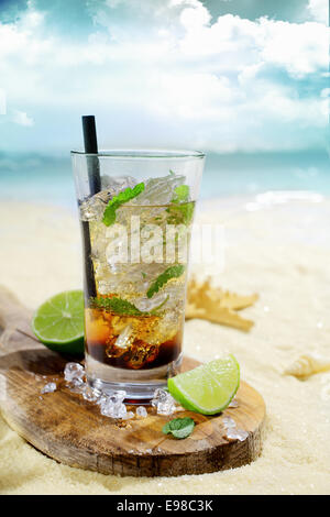 Erfrischenden Eistee Rum cocktail in ein Longdrinkglas auf einem Holzbrett mit in Scheiben geschnittenen Zitrone auf dem goldenen Sand von einem idyllischen tropischen Strand serviert Stockfoto