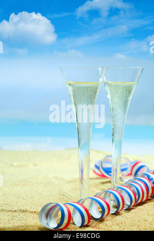 Sektgläser im eleganten geriffelten Gläsern und einem bunten roten, weißen und blauen gezwirbelten Partei Streamer stehen auf dem goldenen Sand von einem tropischen Strand mit einem azurblauen Ozean Blau auf einen Sommerurlaub. Schauen Sie sich mein Portfolio für eine ganze Reihe von Cocktails. Stockfoto