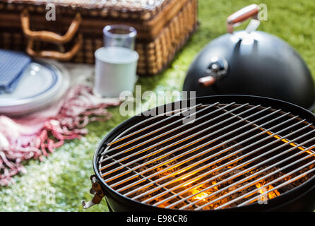 Ein brennendes heißes Feuer in einem tragbaren Grill mit einem leeren Grill und ein Korb Picknickkorb sichtbar auf einem grünen Rasen hinter Nahaufnahme Stockfoto
