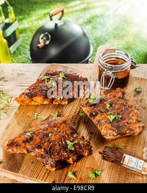 Vorbereitung von Rippen für ein BBQ mit Bohnenkraut Heften Sauce und fein gehackte frische Kräuter mit drei Portionen auf einem Tisch mit einem Glas Marinade und einer Bürste Stockfoto