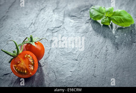 Frisch halbierte Tomaten und frischem Basilikum, angeordnet den gegenüberliegenden Ecken des Rahmens auf eine dunkle strukturierte Oberfläche in einem flachen Winkel Stockfoto