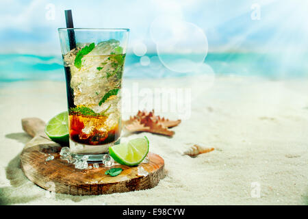 Erfrischender Eistee Rum, Limette und Minze Mojito cocktail stehend in einem hohen Glas auf einem alten Holzbrett auf dem goldenen Sand von einem idyllischen tropischen Strand, einen Sommerurlaub zu feiern Stockfoto