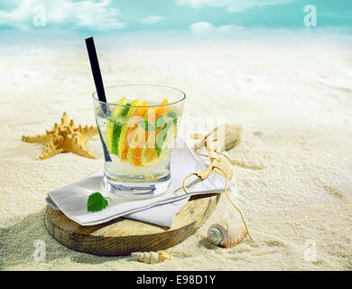 Glas erfrischenden gekühltes Gin oder Wodka-cocktail mit Scheiben von Zitrone, Limette und Orange auf einem tropischen Strand mit einer Muschel und Seesterne auf goldenem sand Stockfoto