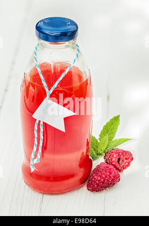 Hausgemachten frischen Himbeersaft in einer Glasflasche aus frisch gepressten Früchten zusammen mit zwei frischen Beeren auf einem rustikalen weißen hölzernen Hintergrund gemacht Stockfoto