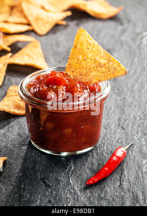 Glas heiße würzige Tomaten-Salsa mit Nachos oder Mais-Tortillas mit einer roten Cayennepfeffer Chili auf dunklem Schiefer Hintergrund Stockfoto