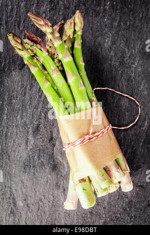 Frische grüne Spargelstangen gebunden in einen braunen Papier-Wrapper für eine gesunde vegetarische Küche liegen auf einem grauen Schieferoberfläche, Draufsicht Stockfoto