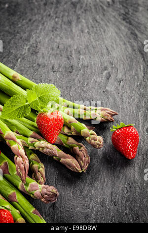 Frische ungekochte grünen Spargel, Erdbeeren und Pfefferminz auf einem grauen Schiefer Oberflächenstruktur mit Exemplar in Hochformat Stockfoto
