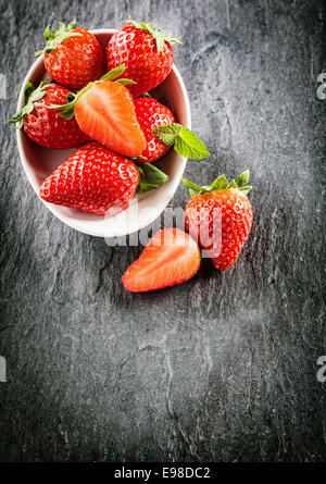 Ausschank von süßen saftigen Erdbeeren in einer weißen Schüssel garniert mit Minze mit einer halbierten Frucht auf die Schieferoberfläche neben, Stockfoto