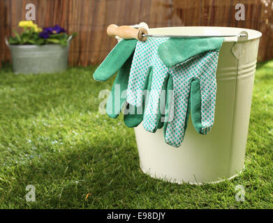 Grüne Textile Gartenhandschuhe hängen über den Rand des einen Blecheimer stehen auf einem grünen Rasen im Sommersonnenschein, Konzeptbild Stockfoto