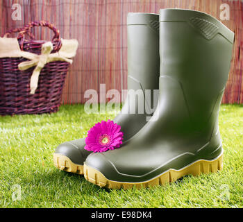 Gummistiefel mit einem rosa Gerbera Daisy stehen auf einem gepflegten grünen Rasen vor einem rustikalen Weidenkorb in einem Garten- und gesunden Lifestyle-Konzept Stockfoto