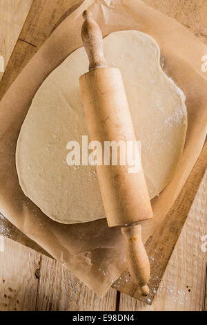 Hausgemachte Pizzabacken in einer Landhausküche mit eine Draufsicht des ausgerollten Teig für den Boden mit einem hölzernen Nudelholz auf Ofen Papier auf einem alten hölzernen Schneidebrett Stockfoto