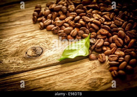 Hintergrund der braunen gerösteten Kaffeebohnen auf alte Grunge rustikale verwitterten Treibholz Dielen mit einem frischen grünen Blatt, Exemplar und Vignettierung Stockfoto