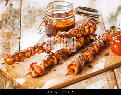 Marinierte heiße würzige Kebabs auf einen Sommer BBQ mit Cherry-Tomaten gegrillt und serviert auf einer alten hölzernen hacken Baird auf einem rustikalen verwitterten Picknick-Tisch mit einem Glas von Heften Gewürz Stockfoto