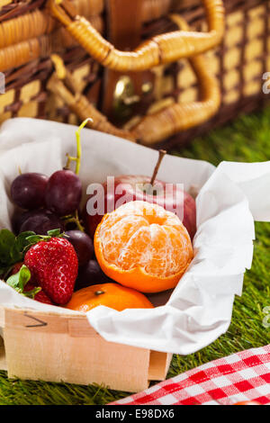 Sommer Picknickkorb auf dem grünen Rasen mit einer Box gefüllt mit gesunden frischen Früchten, einschließlich einer geschälten Nektarine, Erdbeeren, Trauben und einen Apfel für einen leckeren snack Stockfoto