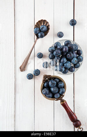 Draufsicht der Blaubeeren oder Heidelbeeren in einer antiken Teesieb, Glasschale und alte Löffel über einem weißen Holztisch Stockfoto