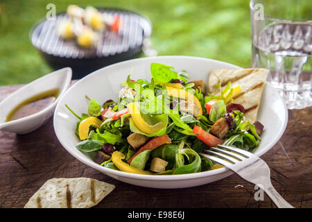 Leckere Kräutersalat mit frischen grünen Blätter Rucola, Paprika und Tomaten serviert mit einer Öl-Dressing auf einem Sommer-Picknick-Tisch im Garten Stockfoto