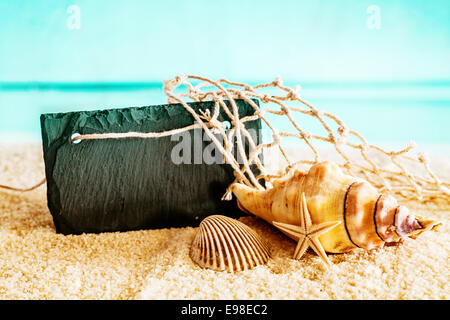 Schöne tropische nautischen Stillleben mit einem leeren schwarzen Schild Exemplar auf goldenen Meer Sand auf einem idyllischen Strand zusammen mit einer Muschel, Herzmuschel und Seesterne mit einem Stück Fischernetz stehen Stockfoto