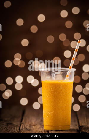 Kunststoff-Glas lecker erfrischend kühlen tropischen orange Mango Smoothie stehend auf einem hölzernen Theke mit Blick auf einen funkelnden Bokeh Party Lichter mit Exemplar Stockfoto