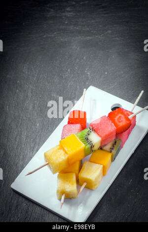 Trio of fresh fruit kebabs with cubes of strawberry, melon, watermelon, kiwifruit, and pineapple on wooden skewers served on a plate, high angle view on slate with copyspace Stockfoto