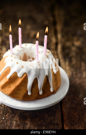 Erhöhte Ansicht einer leckeren frisch gebackenen Vanille Geburtstagstorte mit tropfenden weißen Puderzucker glasiert und Perlen und dekoriert mit vier beleuchteten Party Kerzen auf einem rustikalen Holztisch mit Exemplar Stockfoto