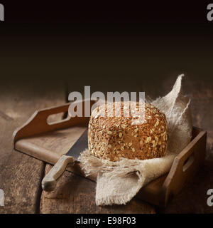 Leckere Vollkorn Landbrot für einen gesunden Snack oder Beilage zu einer Mahlzeit serviert auf ein altes rustikales Holztablett mit einem Brotmesser mit dunklen Exemplar hinter Stockfoto