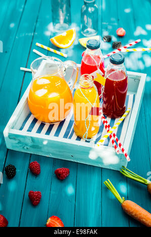 Frische gesunde hausgemachte Fruchtsäfte gequetscht aus Zitrusfrüchten, Himbeeren Erdbeeren und Möhren stehend auf einem Holztablett in Glasflasche auf einem türkisfarbenen Picknicktisch im gefleckten Sommer Sonne Stockfoto