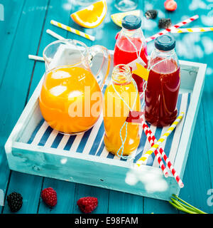 Flaschen von frisch gepressten Orange und Berry Saft stehend auf einem Holztablett auf einem bunten Türkis Blau Picknicktisch im gefleckten Sommersonne Stockfoto