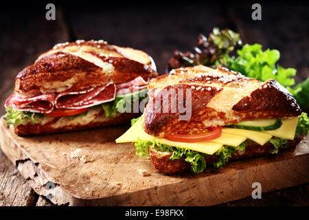 Knusprig braun frisch gebackene Lauge Brötchen mit Käse, pikante Salami Wurst, Salat, Tomaten und Gurken auf eine alte Grunge rustikale Schneidbrett aus Holz Stockfoto