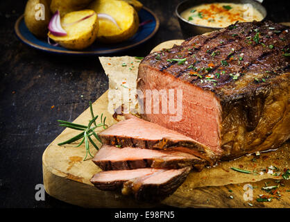 Geschnitzte selten Roastbeef gewürzt mit frischem Rosmarin, Gewürzen und Kräutern auf einem alten hölzernen Schneidebrett in einer Landhausküche Stockfoto