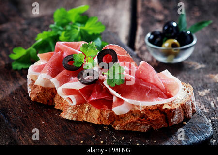 Leckere Schinken oder deutschen Schinken Sandwich mit dünnen Scheiben von Gourmet-Fleisch auf Roggenbrot serviert und garniert mit in Scheiben geschnittenen Oliven und frischen Kräutern in eine rustikale Landhausküche Stockfoto