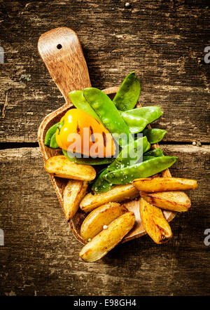 Beilage lecker gekochtes Gemüse serviert auf einem Holzspatel mit gerösteten süßen Paprika, grüne Zuckererbsen Erbsen und Kartoffelspalten auf einem rustikalen Grunge Holz Hintergrund Stockfoto