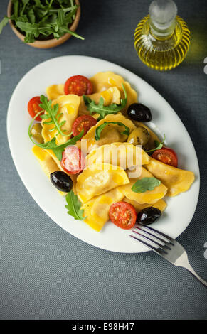 Italienische Ravioli Pasta mit Oliven, Tomaten und Rucola Blätter mit einem Dekanter von nativem Olivenöl als Dressing, obenliegende Ansicht auf einer ovalen Platte serviert Stockfoto