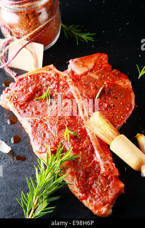 Rohe Porterhouse-Steak mit pikanten Gewürzen und ein Zweig frischer Rosmarin neben ein Glas das pulverisierte Gewürz mit einem leeren Etikett während der Zubereitung von Fleisch zum Grillen in eine rustikale Küche Stockfoto