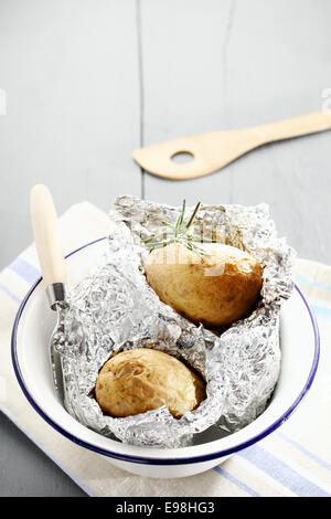 Frische heiße Ofenkartoffeln mit Rosmarin noch eingebettet in ihre Folie-Wrapper in ein Metall Becken auf einem weiß lackierten hölzernen Picknicktisch mit Exemplar Stockfoto