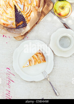 Stück leckeren frisch gebackenen Apfelkuchen serviert auf einem Teller mit einer leeren Tasse und Untertasse für Tee oder Kaffee neben den ganzen Kuchen auf ein weißes Tuch von oben gesehen Stockfoto