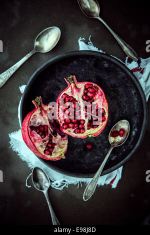 Blick von oben auf einen frischen halbierten Granatapfel auf einer Platte zeigt die saftige Reifen rote süße Samen mit Löffel zum Essen auf einem dunklen Hintergrund Stockfoto