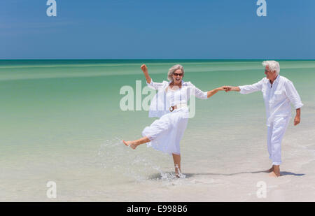 Glückliche senior Mann und Frau paar tanzen, Wasser Händchenhalten & planschen im Meer auf einem einsamen tropischen Strand mit hellen klaren Stockfoto