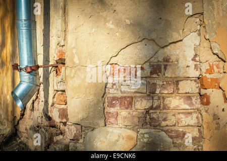 Gelbe Wand der alten Gebäude mit sichtbaren roten Ziegelsteinen geknackt. Einige Schichten von Materialien. Metall Fallrohr hängen. Schatten an der Wand. Stockfoto
