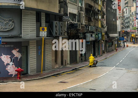 Eine Frau in einem gelben Regenmantel schiebt einen Einkaufswagen in eine leere Straße auf der Insel Hong Kong Stockfoto