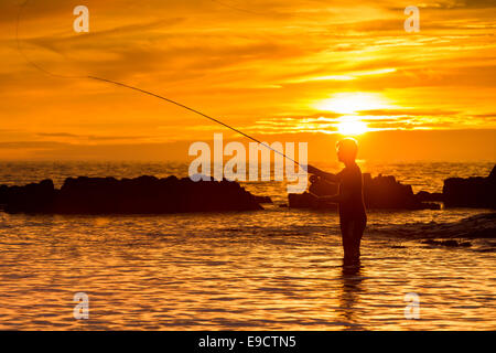 Ein einsamer Angler Fische aus einem felsigen Strand Silhouette gegen die untergehende Sonne Stockfoto