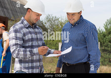 Zwei Architekten Überprüfung Spezifikationen auf einem handheld Dokument, wie sie miteinander reden auf einer Baustelle mit einem Arbeiter im Hintergrund arbeiten. Stockfoto
