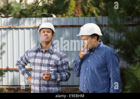 Zwei Ingenieure nehmen einen Rauch brechen in ihre Schutzhelme im Schatten eines Baumes auf einer Baustelle schnaufend auf ihre Zigaretten. Stockfoto