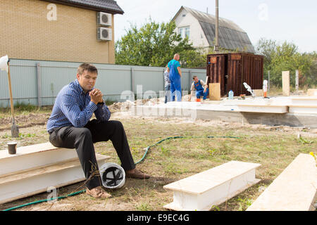 Mittleren Alter Mann Bauingenieur eine Pause mit Helm am Boden. Sitzend auf weißen Balken und etwas zu denken. Stockfoto