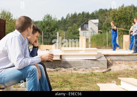 Mittleren Alters männlichen Ingenieure sitzen auf Ecke am Projektstandort mit Bauarbeiter im Hintergrund. Stockfoto
