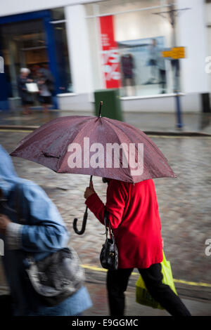 Frau in einer roten Jacke hält einen kastanienbraunen Regenschirm und halten eine gelbe Tragetasche zu Fuß schnell auf einer gepflasterten Straße im Regen Stockfoto