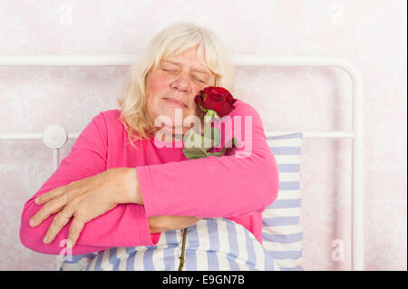 Glückliche Frau in rosa Pyjama sitzen im Bett umarmt eine rose Stockfoto